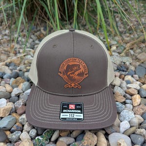 May include: Brown and tan trucker hat with a custom text area and a fishing-themed emblem. The hat features a mesh back, a curved brim, and the Richardson 112 label.