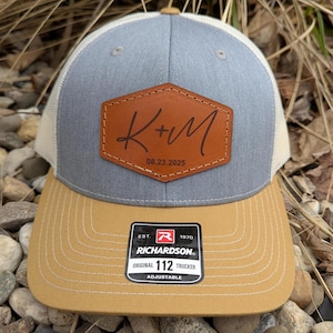 May include: A gray and white trucker hat with a brown leather patch that says "K+M 08.23.2025". The hat is adjustable and has a brown leather patch with the text "EST. 1970 RICHARDSON. ORIGINAL 112 TRUCKER ADJUSTABLE".