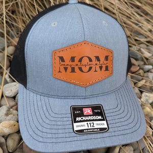 May include: A blue trucker hat with a black mesh back. The front features a brown leather patch with the word "MOM" and the names "Tommy, Taylor, Tyler". The hat has a Richardson 112 Trucker tag.