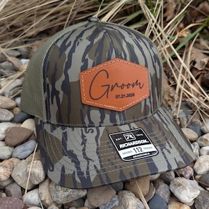 May include: A camouflage trucker hat with a brown leather patch that says "Groom 07.21.2025". The hat is made by Richardson 112.