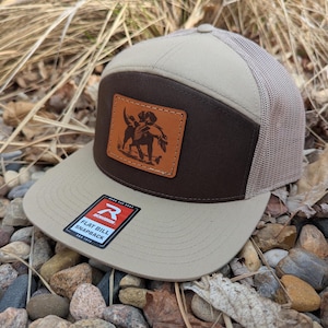 May include: A brown and tan flat bill snapback hat with a brown leather patch featuring a black and white illustration of a dog with a bird in its mouth. The hat has a mesh back and the words "Flat Bill Snapback" are printed on a red tag.
