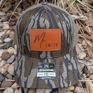 May include: A camouflage trucker hat with a brown leather patch that says "Mr. Smith". The hat is adjustable and has a Richardson 112 label.