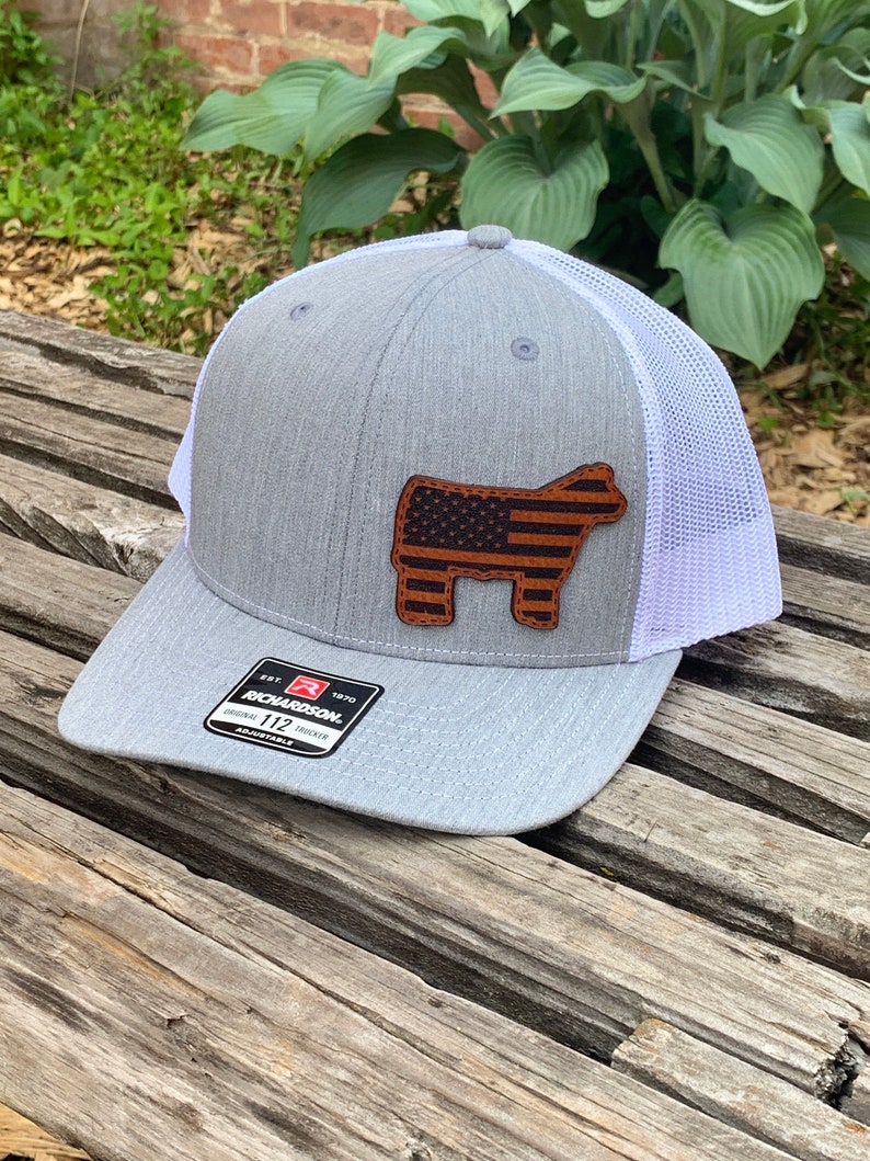 May include: Gray and white trucker hat with a brown leather patch featuring an American flag on a steer silhouette. The hat has a Richardson 112 adjustable label.