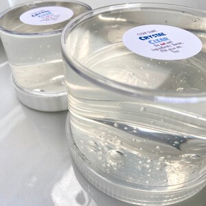 May include: Two clear plastic containers filled with a clear slime. The containers have white lids and labels that say "CLEAR SLIME CRYSTAL CLEAR Do not eat! Ingredients: Borax, PVA glue etc."