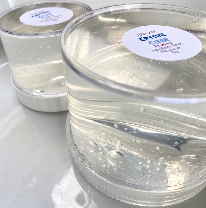 May include: Two clear plastic containers filled with a clear slime. The containers have white lids and labels that say "CLEAR SLIME CRYSTAL CLEAR Do not eat! Ingredients: Borax, PVA glue etc."