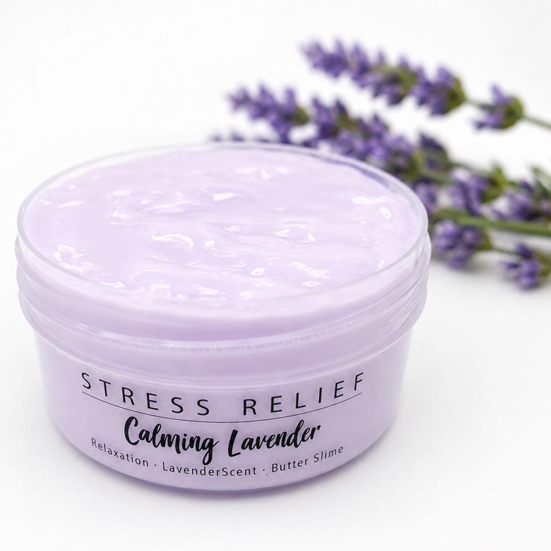 May include: A round, clear plastic container filled with light purple butter slime. The label reads STRESS RELIEF Calming Lavender, Relaxation, LavenderScent, Butter Slime. A sprig of purple lavender flowers is positioned behind the container.