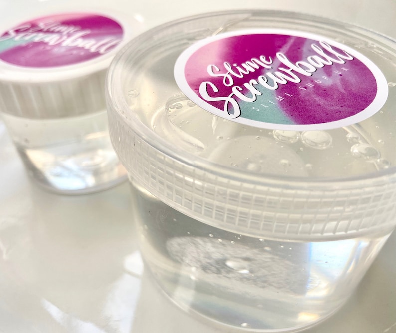 May include: Two clear plastic containers filled with clear slime. The containers have white lids with a pink and blue sticker that says "Slime Screwball Slime Shop".