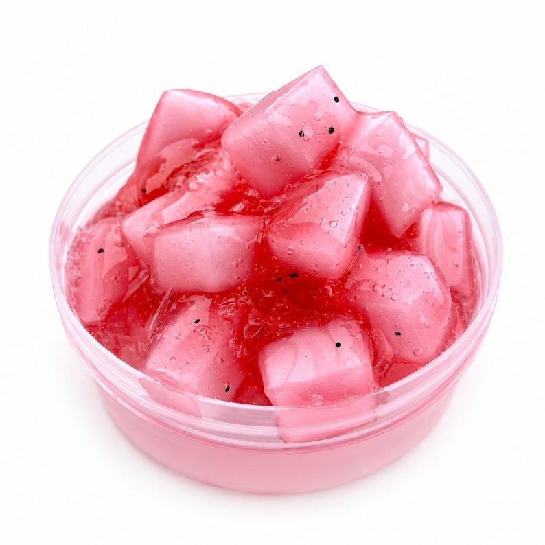 May include: A round, clear plastic container filled with a translucent, vibrant pink slime base. The slime is embedded with numerous semi-translucent, light pink cube-shaped pieces that resemble fruit chunks, each featuring small black speckles.