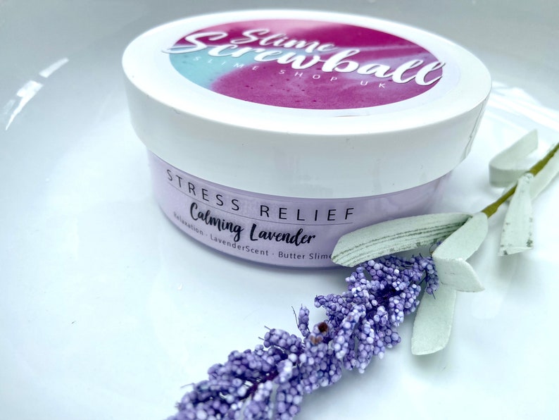 May include: A white jar of "Slime Screwball" butter slime with a lavender scent, labeled "Stress Relief" and "Calming Lavender." A sprig of lavender and a white surface complete the image.