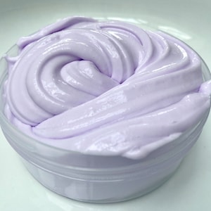 May include: A container filled with lavender-colored slime. The slime has a smooth, glossy texture and is swirled in a circular pattern. The clear plastic container sits on a white surface.