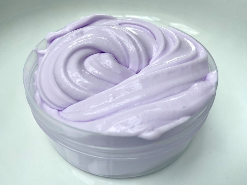 May include: A container filled with lavender-colored slime. The slime has a smooth, glossy texture and is swirled in a circular pattern. The clear plastic container sits on a white surface.
