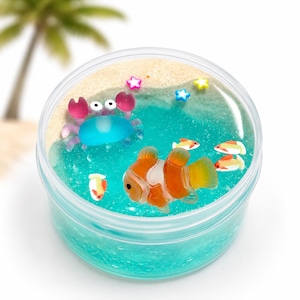 May include: A clear round container filled with blue textured slime representing water and a sandy beach area. The slime features small plastic decorations including a blue crab, an orange and white clownfish, three smaller fish, and three colourful star-shaped charms.