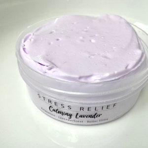 May include: A small, clear plastic container filled with light purple butter slime. The container has a white label with the words "STRESS RELIEF Calming Lavender" printed on it. The slime has a smooth, creamy texture.