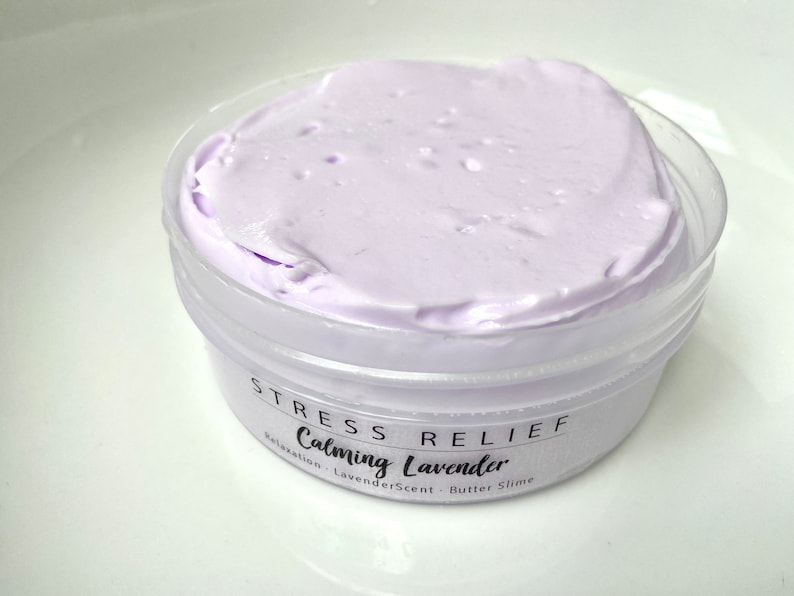 May include: A small, clear plastic container filled with light purple butter slime. The container has a white label with the words "STRESS RELIEF Calming Lavender" printed on it. The slime has a smooth, creamy texture.