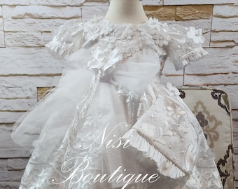 May include: White lace baby dress with a satin bow and a matching bonnet. The dress has a scalloped lace trim and is decorated with white flowers. The bonnet has a ruffled edge and is tied with a satin ribbon.