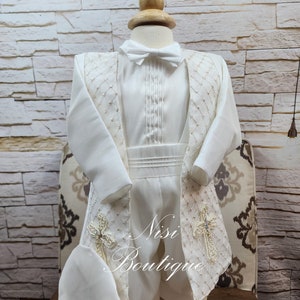 May include: A white christening outfit for a baby boy, featuring a white shirt with a bow tie, a white vest with a cross design, and white pants. The outfit is decorated with pearl-like embellishments and a white hat.