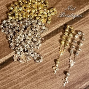 Pode incluir: Um close-up de dois conjuntos de rosários. Um conjunto é dourado e o outro é prateado. Ambos os conjuntos têm uma cruz e um amuleto no final. O texto "Nisi Boutique" é visível na imagem.