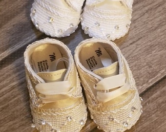 Könnte beinhalten: Ein Paar weiße Babyschuhe mit Spitzendetails und Strasssteinen. Die Schuhe haben ein Satinband und sind mit "Mi" beschriftet.
