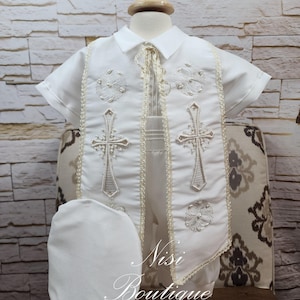 May include: A white baptism outfit for a baby, including a short-sleeved shirt, vest, shorts, and a matching hat. The vest features embroidered crosses and decorative trim. The outfit is displayed on a mannequin.