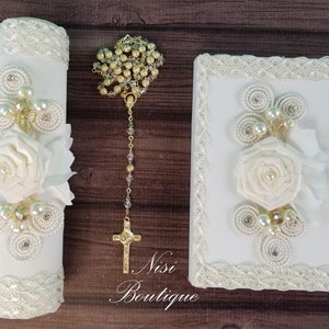 May include: White baptism candle with a white satin rose and pearl embellishments, a white rosary with gold beads and a white prayer book with a white satin rose and pearl embellishments.