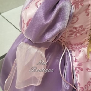 May include: A purple and pink satin dress with a puffed sleeve and a lace trim. The dress has a lace-up back and a pink satin sash.