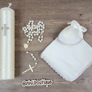 May include: A white satin christening candle with a gold cross, a white pearl rosary, a white lace handkerchief, and a white satin bag with a pearl-like object inside. The items are arranged on a wooden surface. @nisi.boutique