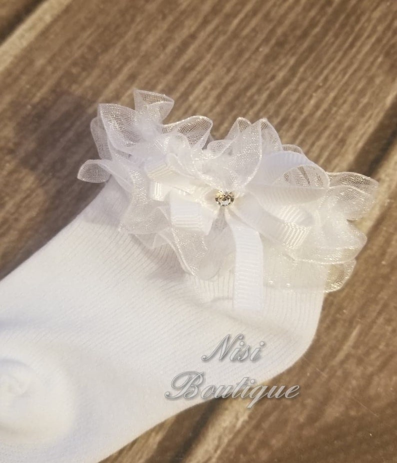 May include: White socks with a white lace ruffle and a small rhinestone embellishment. The socks have the text "Nisi Boutique" on the side.