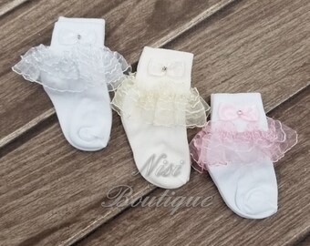 May include: Three pairs of white baby socks with lace ruffles and a jeweled bow on each sock. The socks are in white, cream, and pink.
