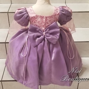 May include: A purple satin dress with a pink satin bodice featuring a floral pattern. The dress has a large bow in the back and is tied with a pink ribbon.