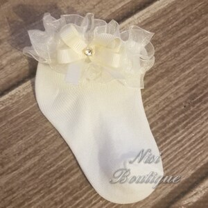May include: White baby socks with a white and ivory lace ruffle and a small rhinestone embellishment. The socks have the text "Nisi Boutique" on the bottom.