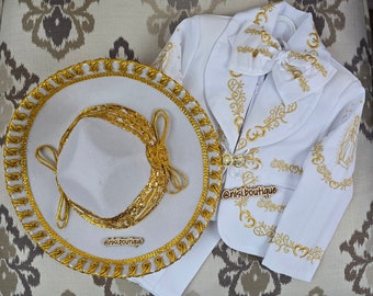 May include: White charro suit with gold embroidery, a white sombrero with a gold trim, and a white bow tie. The suit features a traditional Mexican design with floral patterns and a religious image. The text "@nisi.boutique" is visible on the suit and sombrero.