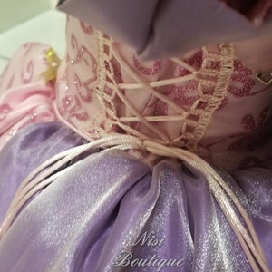 May include: A purple and pink dress with a lace-up back and a tulle skirt. The dress has a sparkly finish and the text "Nisi Boutique" is visible on the skirt.