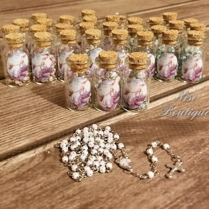 May include: A collection of small glass bottles filled with white beads and a religious image. The bottles have cork stoppers and are arranged in rows on a wooden surface. A white rosary with a silver cross is also pictured.