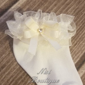 May include: White baby socks with a white and ivory bow and a rhinestone in the center. The bow is made of tulle and has a ruffled edge. The socks have a Nisi Boutique logo on the side.