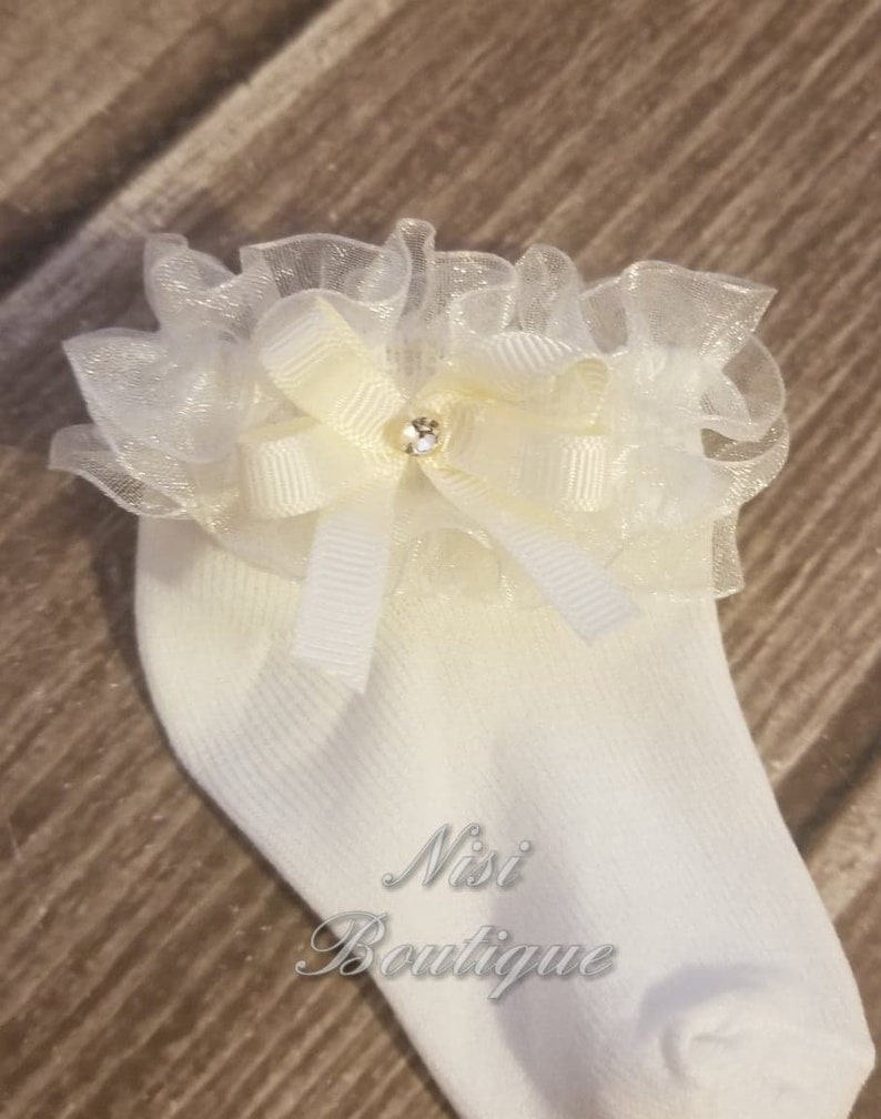 May include: White baby socks with a white and ivory bow and a rhinestone in the center. The bow is made of tulle and has a ruffled edge. The socks have a Nisi Boutique logo on the side.