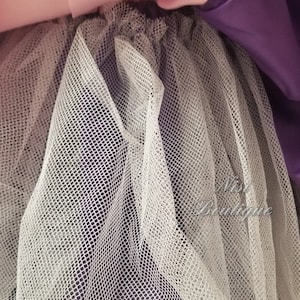 May include: Close-up of a white tulle fabric layered over a purple satin fabric. The tulle is a fine mesh with a delicate texture.