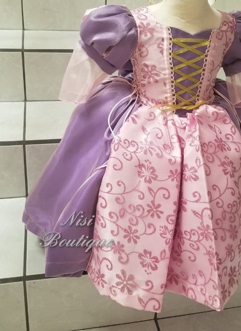 May include: A purple and pink princess dress with a gold ribbon and lace up back. The dress has a floral pattern and is made of satin.