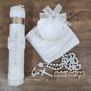 May include: White baptism set with a white candle, a white seashell, a white handkerchief, and a white rosary with a silver cross. The candle has a white lace design and a silver cross. The seashell has a white bow with a silver rhinestone. The handkerchief has a white lace trim. The rosary has white beads and a silver cross. The set is on a wooden surface. @nisi.boutique