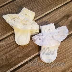 May include: Two pairs of white baby socks with lace ruffles and a satin bow. The socks are on a wooden surface.