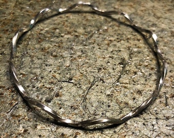 May include: A silver, wavy bangle bracelet. The bracelet is made of a single piece of metal that is twisted and shaped into a circle.