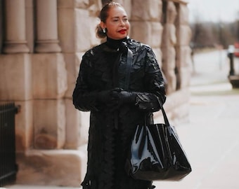 May include: A woman wearing a black, long-sleeved,  layered coat with a black tote bag. She is wearing black heels and has red lipstick.