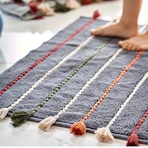 Puede incluir: Una alfombra de baño gris con rayas de colores y borlas. La alfombra tiene una superficie texturizada y está hecha de algodón.