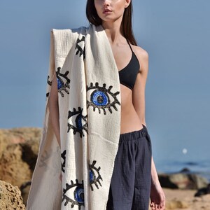 May include: A white cotton beach towel with a fringe border and a pattern of blue and black evil eyes. The towel is draped over a person's shoulders.