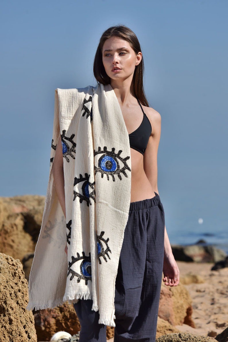 May include: A white cotton beach towel with a fringe border and a pattern of blue and black evil eyes. The towel is draped over a person's shoulders.