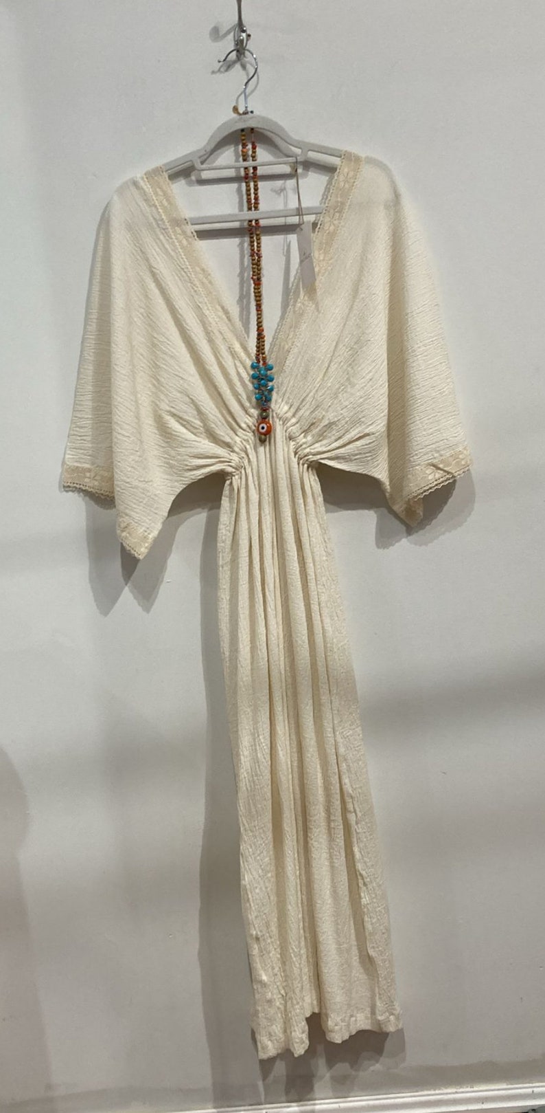 Op de afbeelding: Een witte katoenen jurk met een V-hals en korte mouwen. De jurk heeft een getailleerde taille en een zwierige rok. Het heeft een kralenketting met een blauwe en oranje hanger.