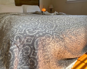 May include: A gray and white patterned throw blanket with a subtle damask design. The blanket is folded on a bed with white pillows.