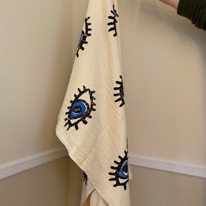 May include: A white cotton throw blanket with a fringe border features a repeating pattern of blue and black stylized eyes.