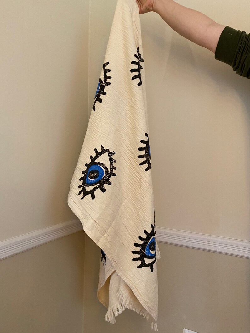 May include: A white cotton throw blanket with a fringe border features a repeating pattern of blue and black stylized eyes.