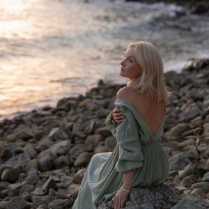 May include: A light green dress with a low back and long sleeves. The dress is worn by a person sitting on a rock near the ocean. The person has blonde hair and is looking towards the horizon. The scene is set at sunset.