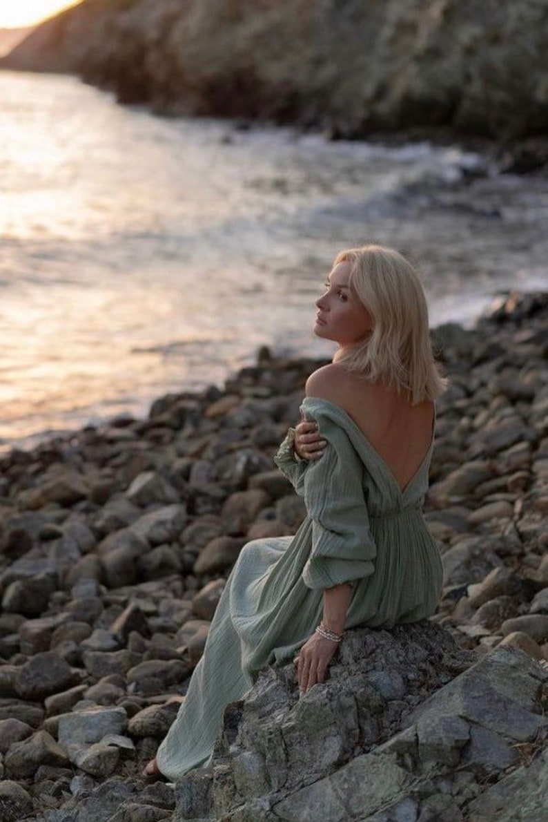May include: A light green dress with a low back and long sleeves. The dress is worn by a person sitting on a rock near the ocean. The person has blonde hair and is looking towards the horizon. The scene is set at sunset.